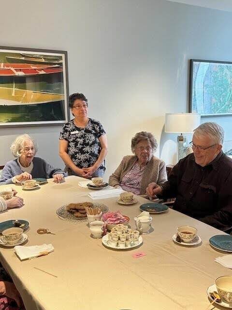 Four seniors and a woman stand and sit around a table set for tea with cups, saucers, snacks, and condiments, in a cozy room with modern art on the walls. This warm gathering highlights dementia activities that promote social engagement and create meaningful moments—perfect as one of many activities for people with dementia at every stage.