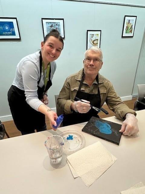 A smiling woman helps an older man with a painting activity at a table, demonstrating engaging Activities for People with Dementia. The man holds a heat tool over a canvas with blue paint, enjoying creative expression as part of meaningful dementia activities. Framed artwork hangs on the wall behind them, showcasing art created through activities for every stage.