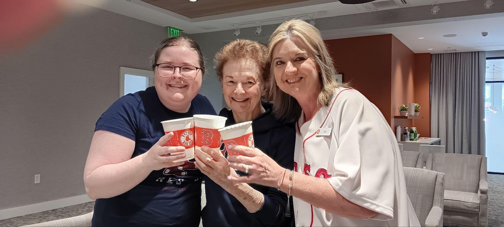 Three smiling women stand indoors, holding up large coffee cups together. The woman on the right is wearing a white Red Sox jersey. The setting appears to be a lounge area with modern decor and comfortable seating, the perfect spot for friends to relax after helping downsize a parent’s home or preparing for a move to memory care.