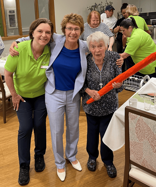 Three women pose and smile in a dining area, one holding an orange foam stick. Other people in green uniforms work in the background, providing attentive memory care. Tables are set with glasses and utensils.