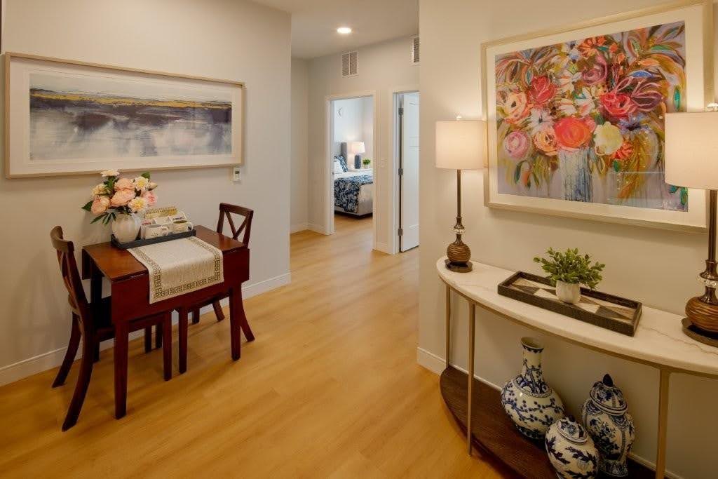 A small dining area with a wooden table and chairs, floral centerpiece, and wall art offers a welcoming space—perfect for those looking to downsize a parent’s home. A hallway leads to a bedroom, while a console table with lamps, vases, and a colorful flower painting sits on the right. Warm lighting and wood floors create an inviting atmosphere ideal for families moving parents or considering memory care needs.
