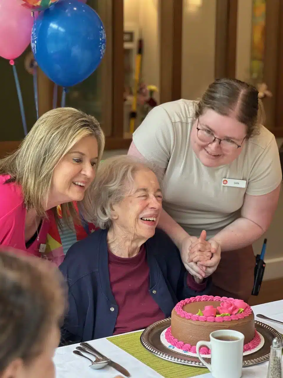 Three women gather around a table with a pink cake. An elderly woman in the center, smiling joyfully, is celebrated by two others—one in pink and one in beige—as blue balloons float nearby, creating a heartwarming moment supported by memory care services.
