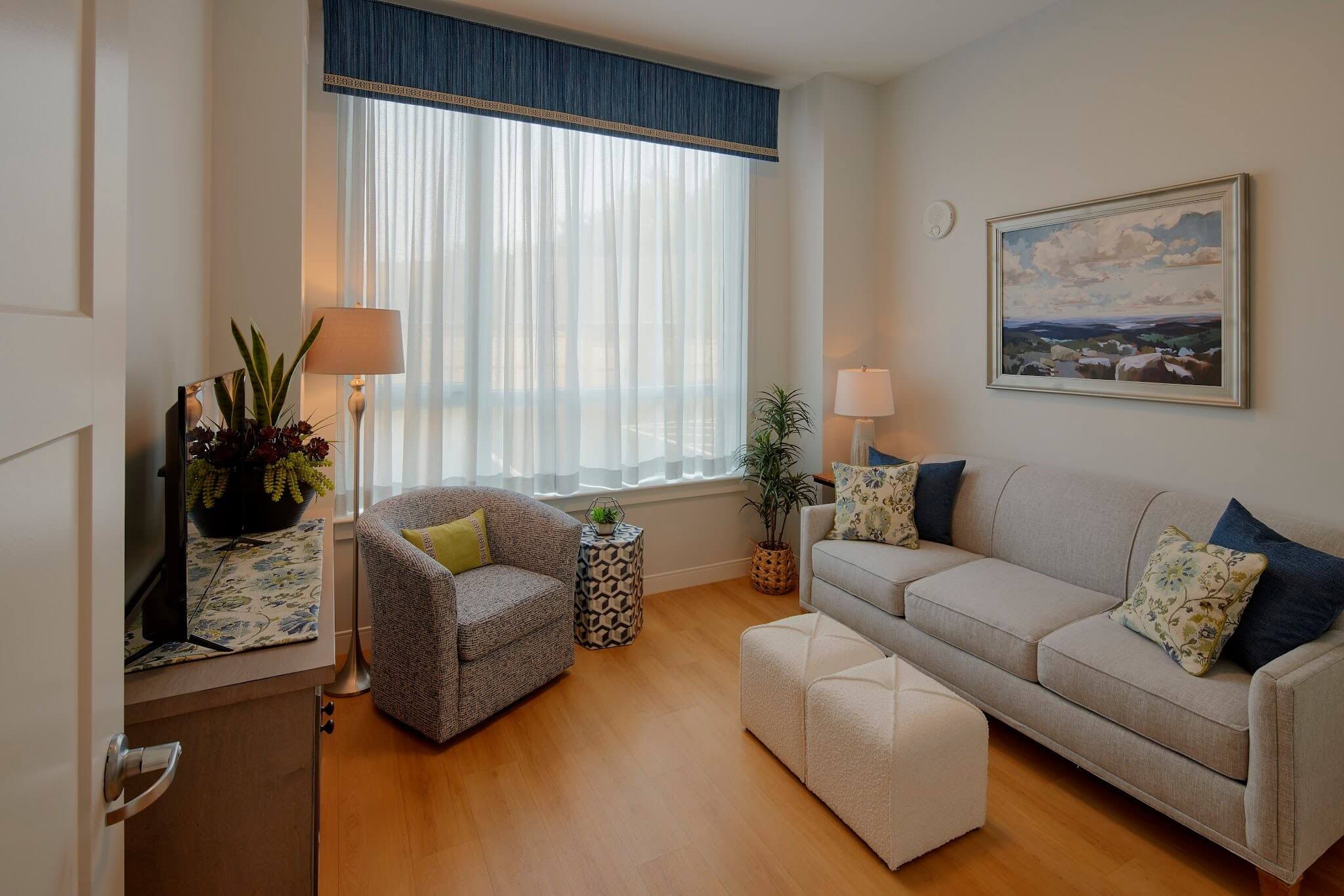 A cozy living room with a gray sofa, an armchair, two white ottomans, plants, a lamp, a TV, and a large window with sheer curtains letting in natural light. A landscape painting hangs on the wall.