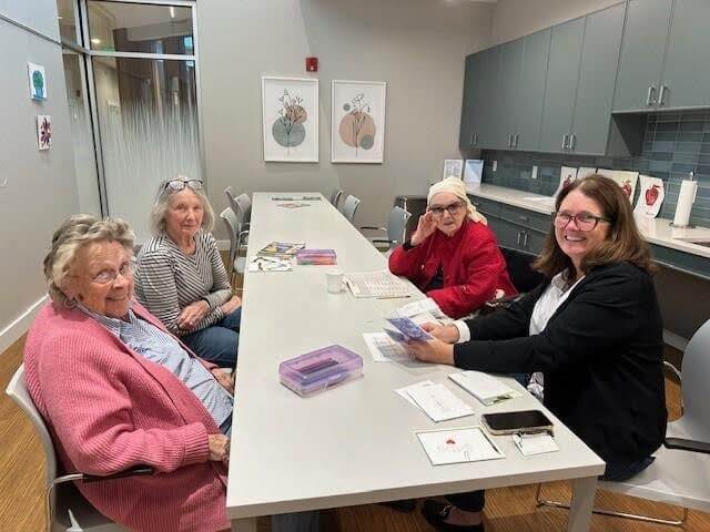 Four women sit around a long white table in a modern room, smiling and engaging with papers and cards. There are shelves and artwork on the walls, creating a friendly, communal atmosphere.