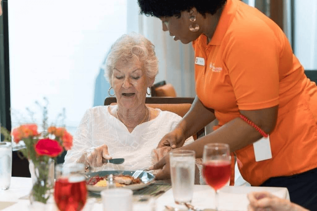 An elderly woman sits at a dining table while a caregiver in an orange shirt helps her with a meal. The table has flowers, water, and red drinks.