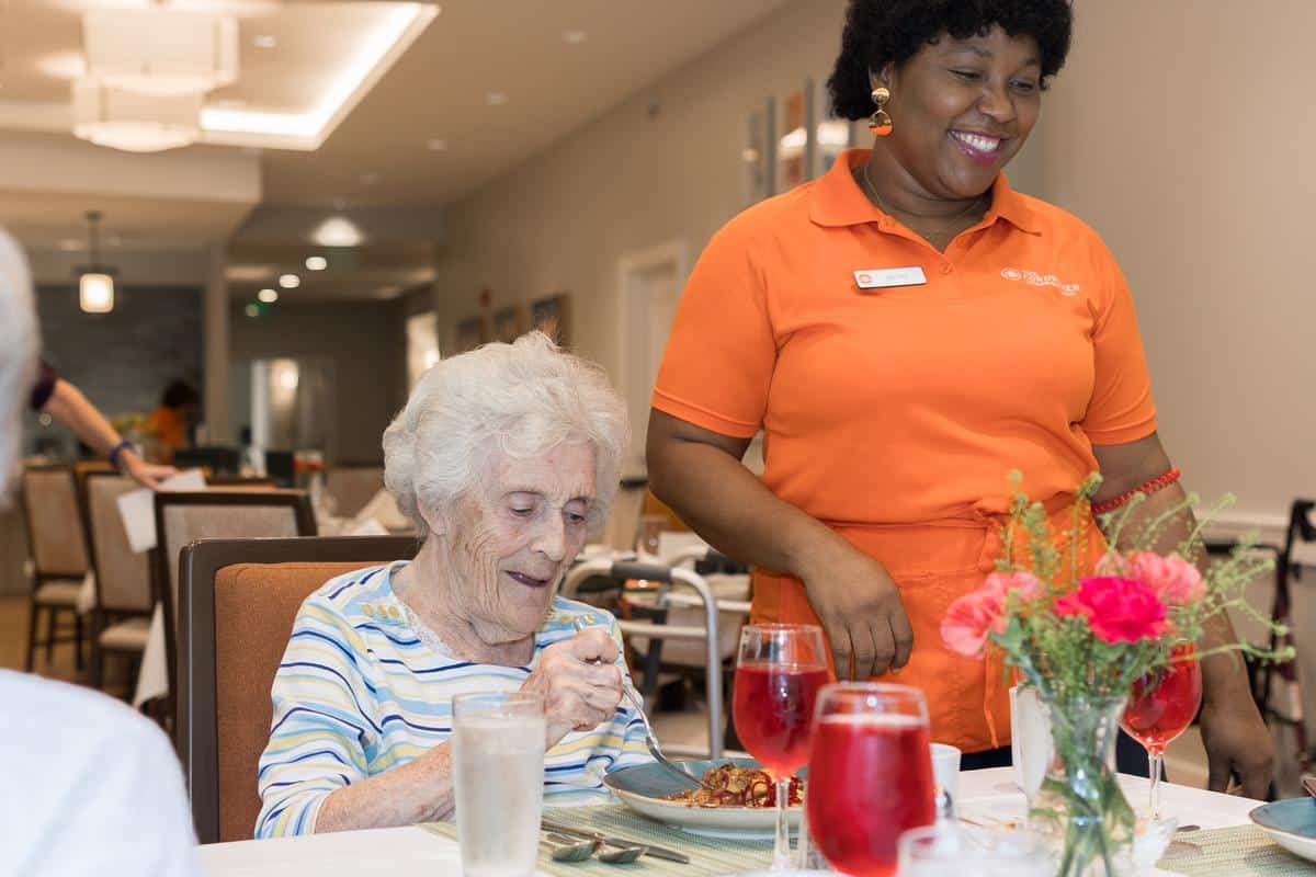 An elderly woman with white hair enjoys a meal at a table, smiling, while a staff member in an orange shirt and name tag stands beside her, also smiling. There are drinks and a flower vase on the table.