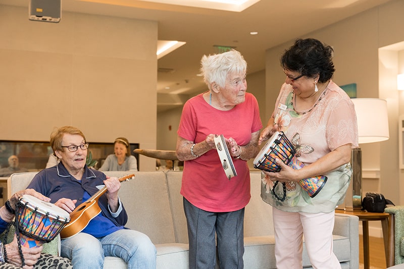 Three elderly women enjoy making Art together indoors; one plays a ukulele, another holds a tambourine and smiles, and the third plays a drum. All appear cheerful and engaged in their creative draft.