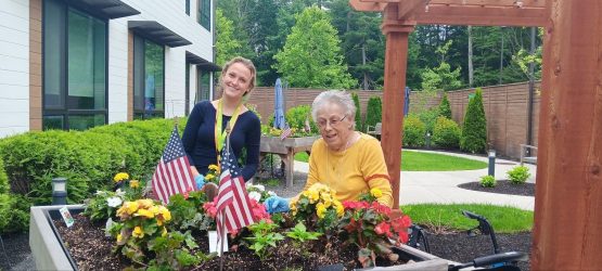 Two women, one younger and one elderly, smile while gardening together at a raised flower bed adorned with colorful flowers and two small American flags, enjoying a sunny day in The Cordwainer’s Dementia Care South Shore MA courtyard.