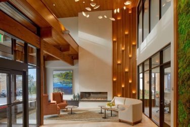 Modern hotel lobby with high ceilings, wood beams, a green living wall, a fireplace, orange chairs, a white couch, coffee tables, and warm lighting. Large windows let in natural light, and contemporary art hangs near the fireplace.