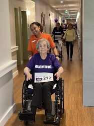 An elderly woman in a wheelchair, wearing a Walk With Me shirt and race number 212, is pushed by a smiling caregiver. Others walk and smile behind them in a brightly lit hallway.
