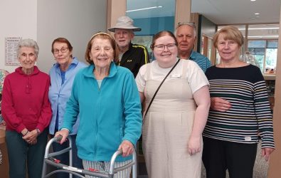 A group of seven adults, including elderly women and men, stand together indoors, smiling at the camera. One woman uses a walker. They appear to be in a welcoming communal or care facility environment.