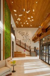 Modern lobby with a green living wall, wooden ceiling, artistic light fixture, staircase with glass and wood railing, and a glass entrance. A curved sofa and small table with yellow flowers are in the foreground.