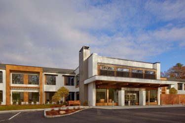 A modern two-story building with large windows, wood and white paneling, and a main entrance canopy, set under a partly cloudy sky. Shrubs and small trees line the front.
