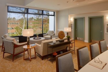 Modern lounge with armchairs, sofas, lamps, and decorative items. Large window shows trees and an office building. Two green doors on the right; a table with chairs and an open notebook in the foreground. Warm, neutral colors throughout.
