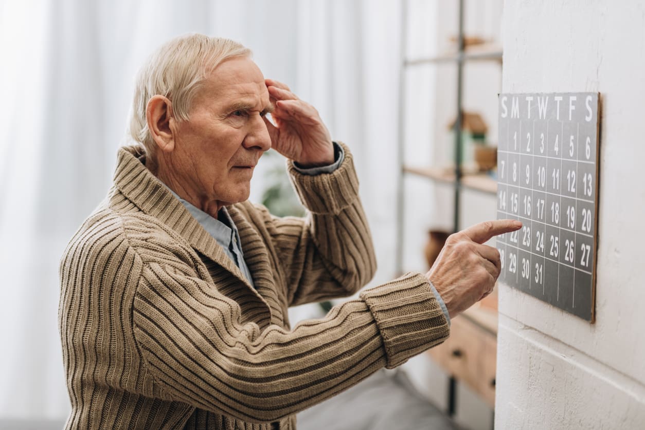 An elderly man in a brown sweater looks confused while touching his head and pointing at a wall calendar, depicting early symptoms of dementia as he tries to remember an important date.