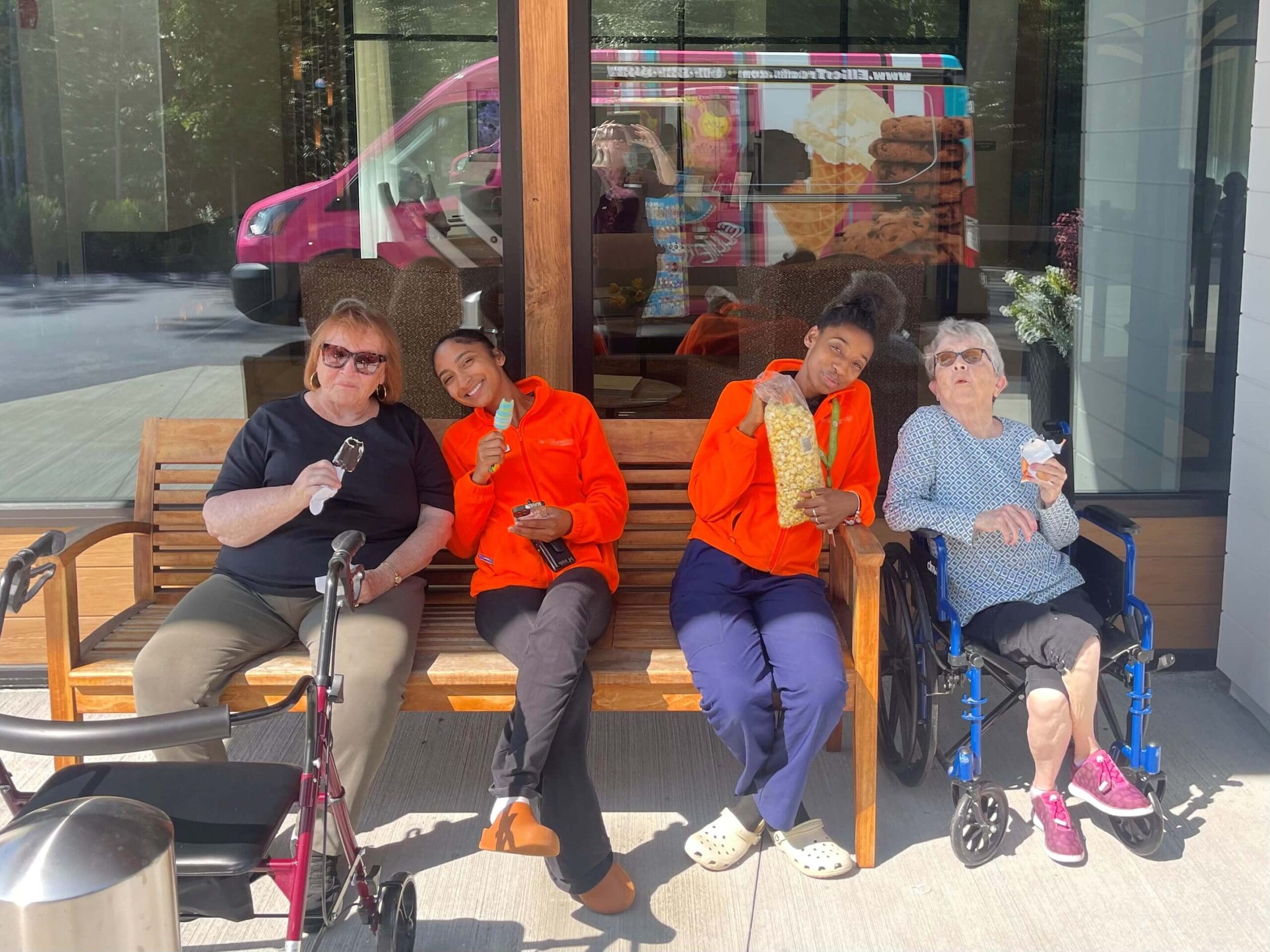 Four women sit on a bench outside, smiling and eating ice cream. Two are in bright orange outfits, one uses a walker, and one is in a wheelchair. Enjoying sweet treats together like this is one of many dementia activities that encourage socialization and joy. A reflection of a pink ice cream truck is visible in the window behind them.