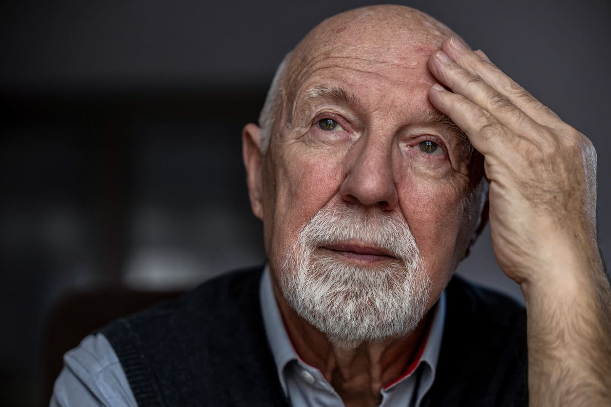 An elderly man with a white beard and bald head rests his hand on his forehead, looking thoughtful and slightly concernedโperhaps experiencing early symptoms of dementia. He is wearing a dark sweater over a collared shirt, with the background softly blurred.