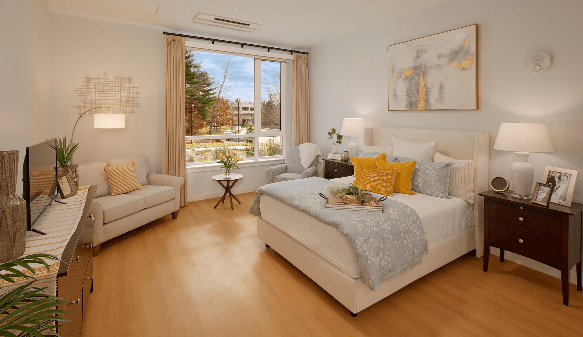 A private resident suite at The Cordwainer with a large window, beige walls, a bed with white and yellow pillows, two nightstands with lamps, a sofa, plants, framed photos, and a light wood floor.