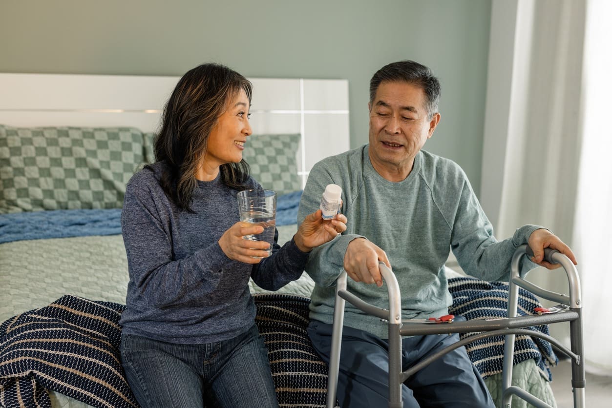 A woman hands a glass of water to an older man with a walker, who is holding a bottle of pills. They are sitting on the edge of a bed, smiling at each other in a brightly lit bedroom.