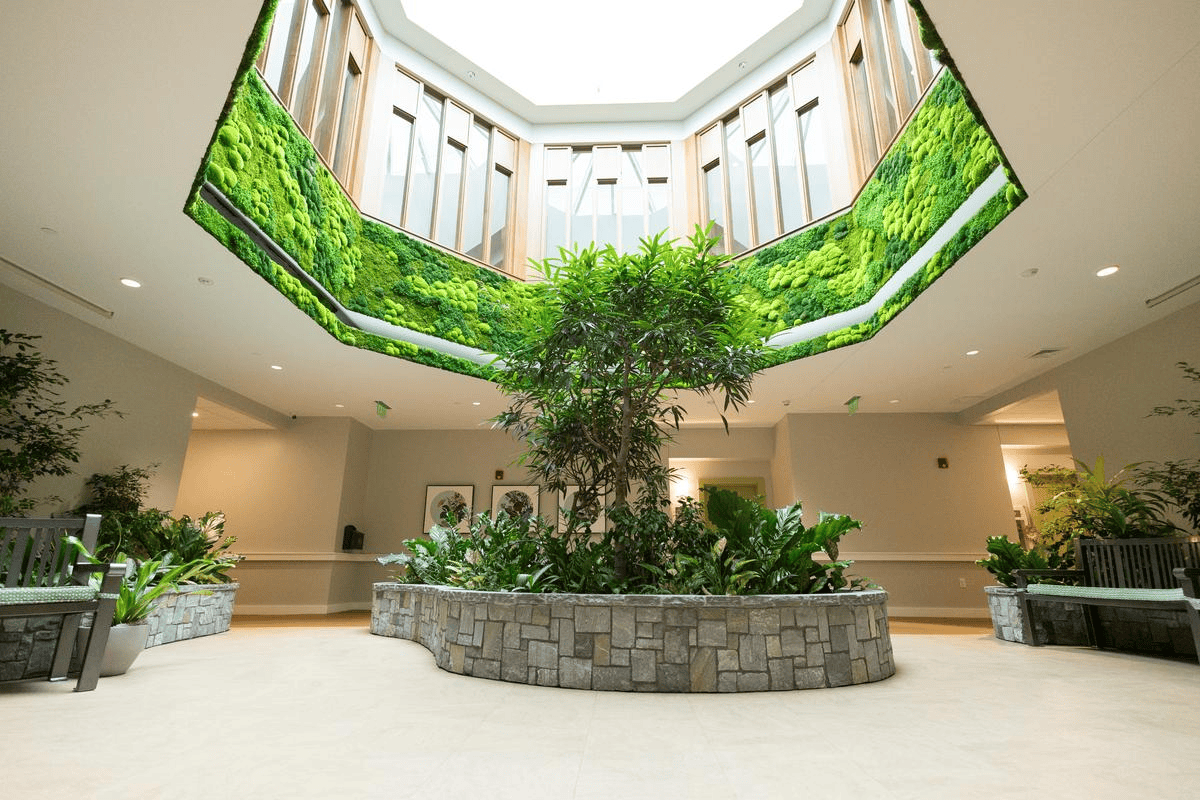 A bright indoor atrium with a central stone planter filled with green plants, surrounded by benches. The upper walls are decorated with panels of vibrant green moss, and a skylight lets in natural light.
