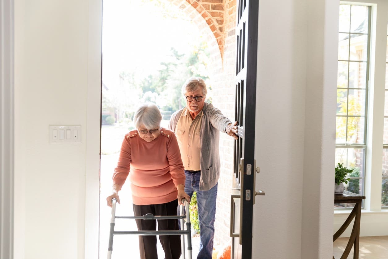 An elderly woman using a walker enters a house with assistance from an elderly man holding the door open for her. Sunlight streams in through the doorway, creating a warm, welcoming atmosphere.