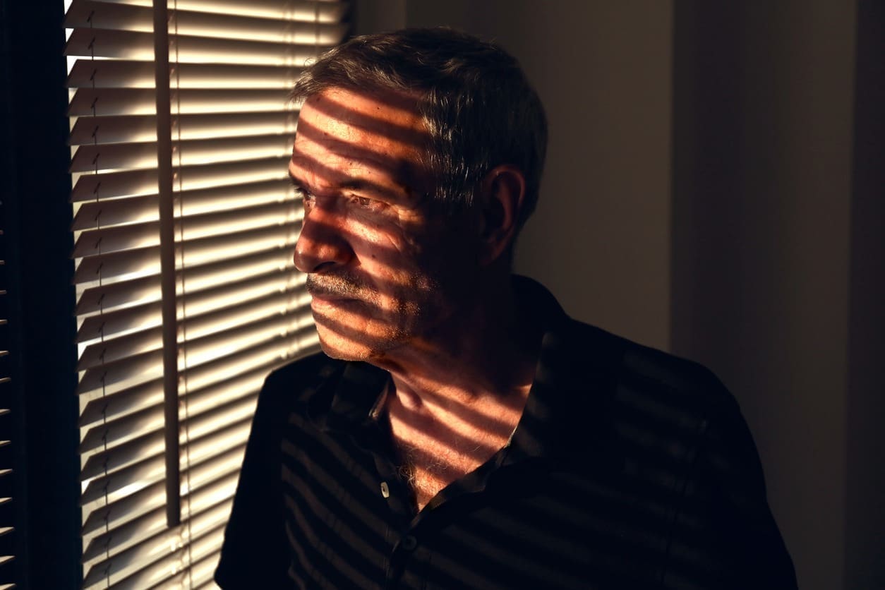 An older man with gray hair and a mustache stands by window blinds, with stripes of sunlight and shadow falling across his face and upper body, creating a dramatic effect.