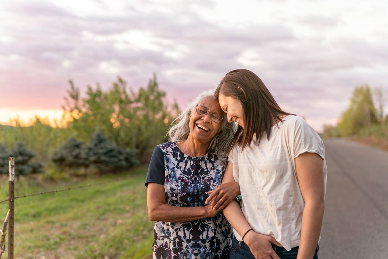 An older woman and a younger woman stand together on a rural road at sunset, smiling and laughing while holding arms. Trees and greenery line the road under a cloudy sky.