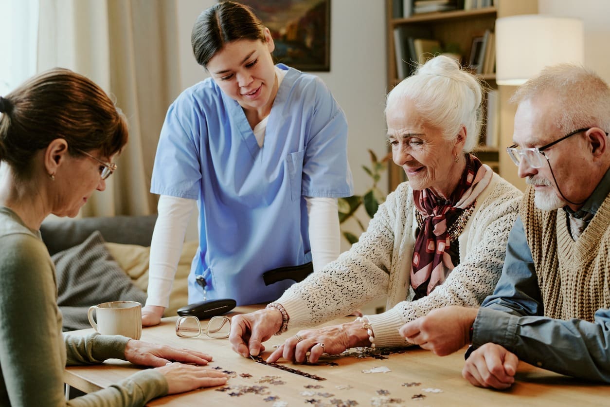 Three elderly people work on a jigsaw puzzle at a table, smiling, with a caregiver in blue scrubs standing nearby and watching them in a cozy living room setting.