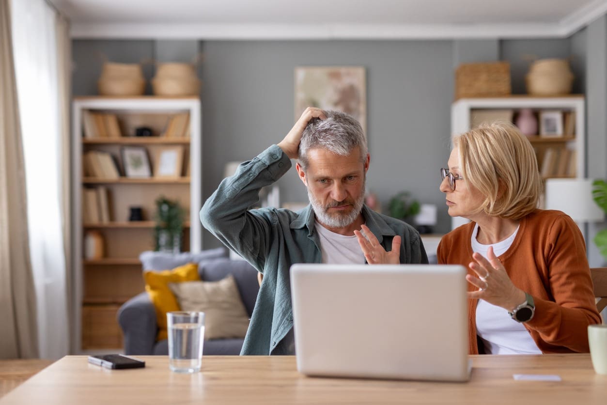 An older man and woman sit at a table with a laptop, looking frustrated and confusedโsigns that could be early symptoms of dementiaโas the man scratches his head and the woman gestures in their cozy, well-lit living room.
