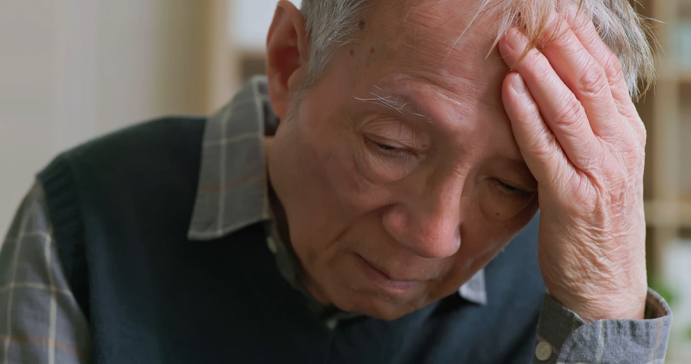 An elderly man with gray hair rests his head in his hand, looking down with a distressed and worried expression. He is wearing a plaid shirt and a dark sweater vest.
