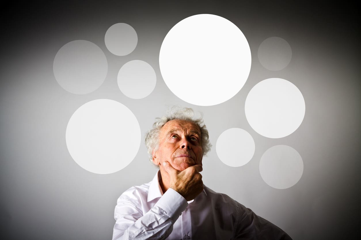 An elderly man with white hair, wearing a white shirt, stands in front of a gray background with large white circles. He looks up thoughtfully, resting his chin on his hand.
