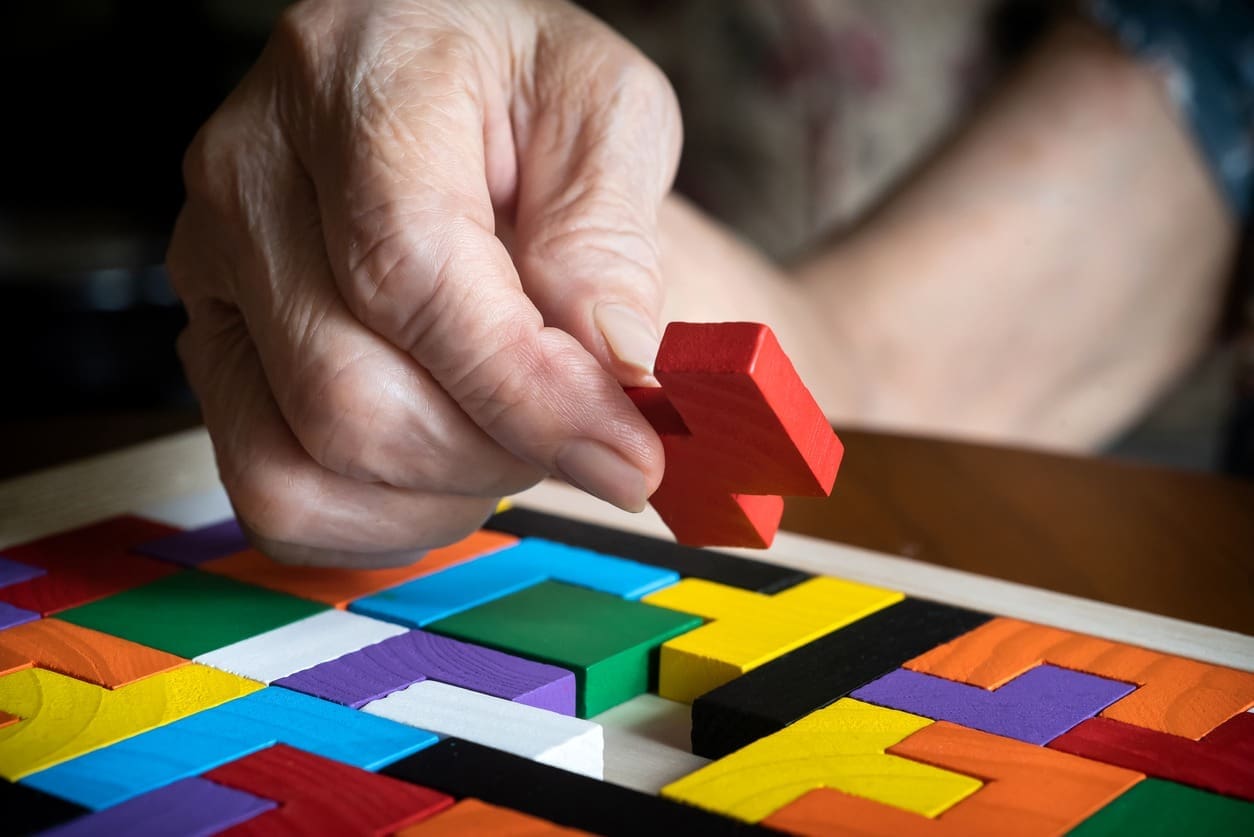 A close-up of an elderly hand placing a red wooden puzzle piece onto a colorful geometric puzzle board.