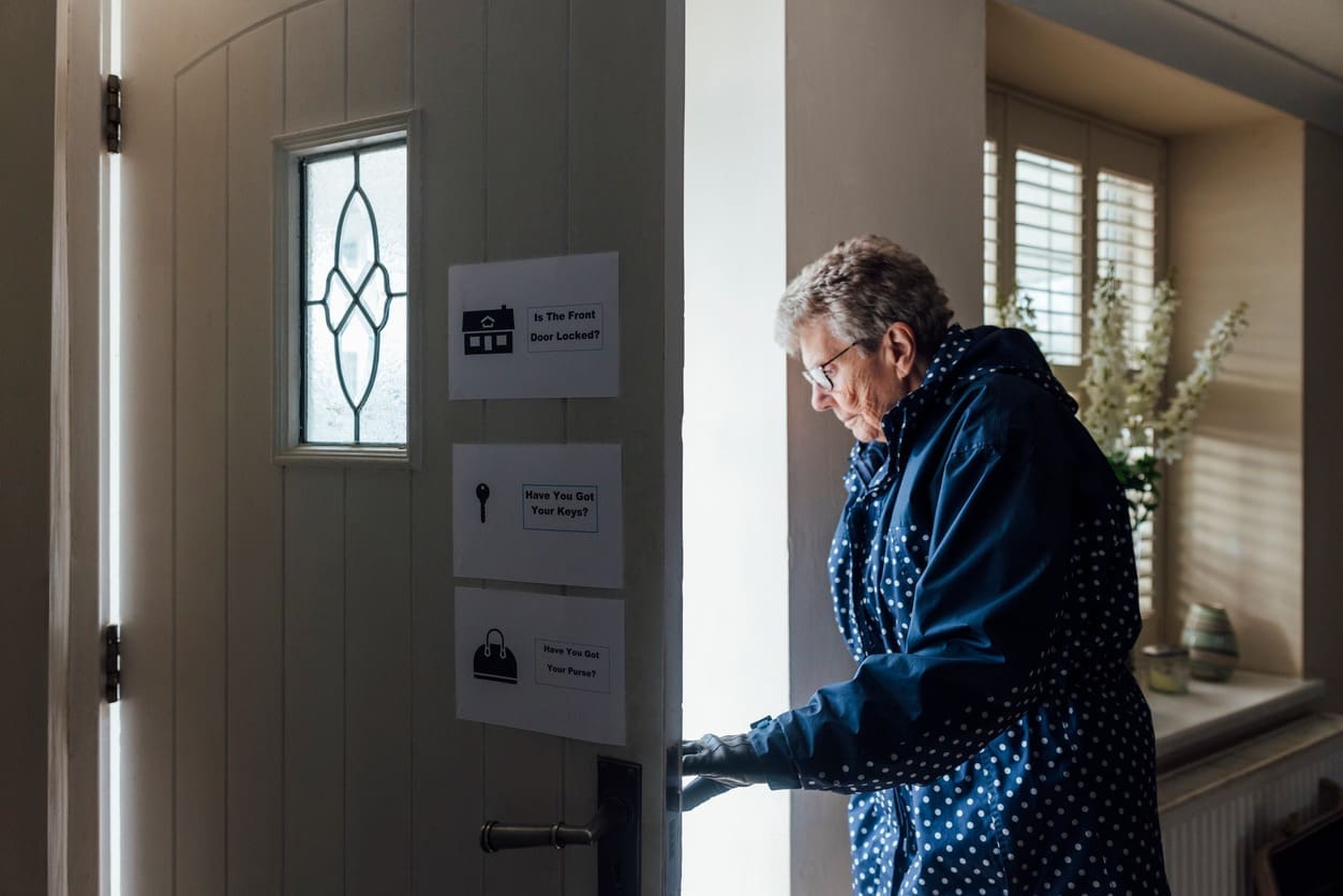 An older woman in a blue polka dot coat, holding keys, locks or unlocks a white front door with reminder notes posted on it—an example of memory care building design fostering supportive environments in a well-lit hallway.