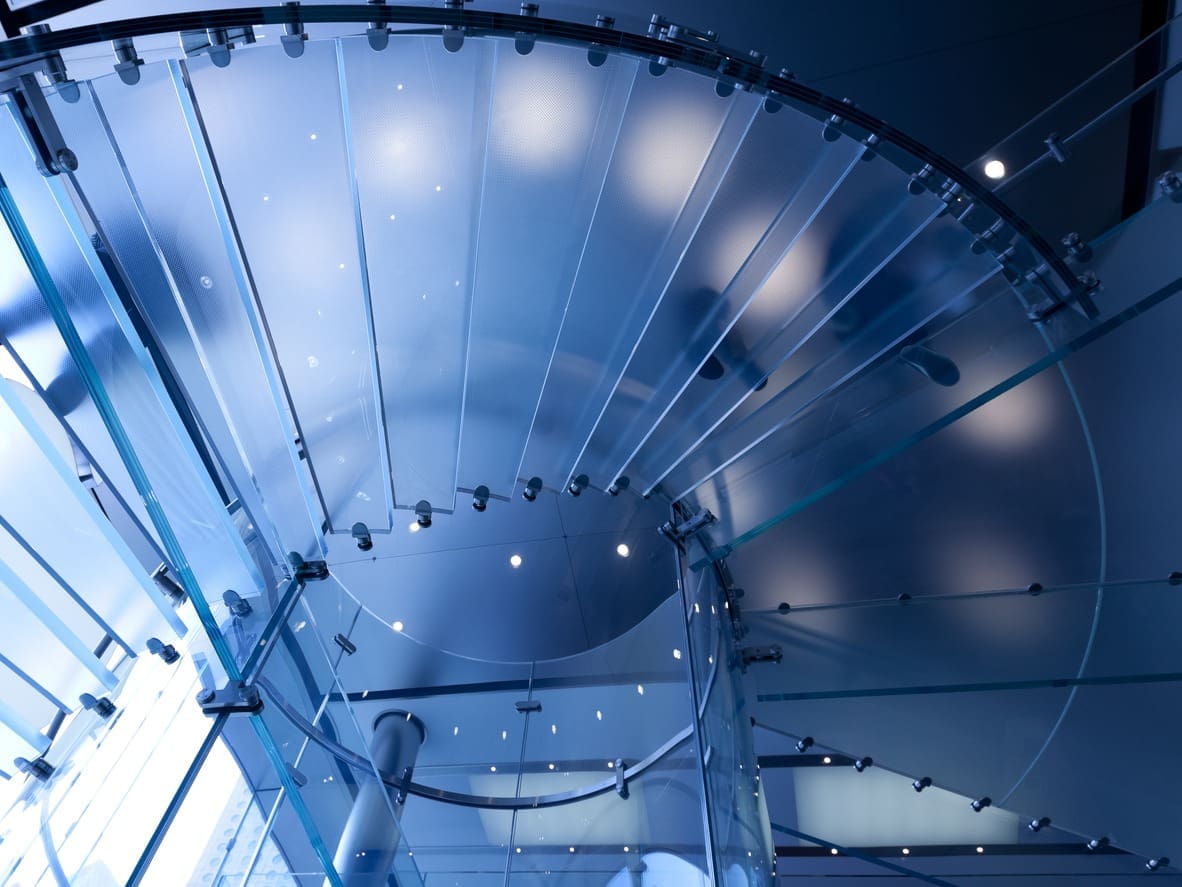 A modern glass spiral staircase viewed from below, with metal supports and spotlights, reflects the principles of human-centered environments. Two blurred silhouettes walk above, casting faint shadows on the translucent steps in a cool blue-toned light.