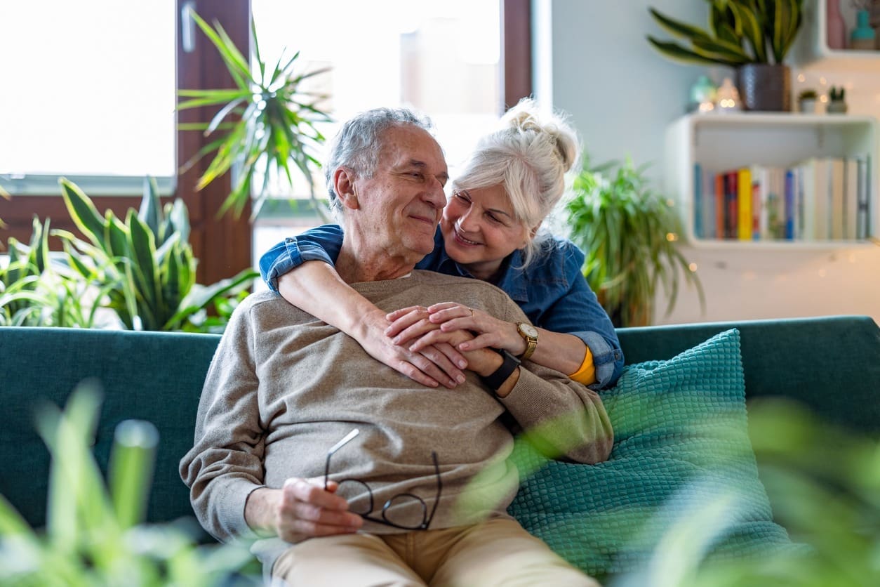 An elderly couple sits on a sofa, smiling lovingly at each other as the woman hugs the man from behind. He holds eyeglasses in his hand. The bright room, filled with plants and books, reflects a supportive environment that nurtures warmth and connection.