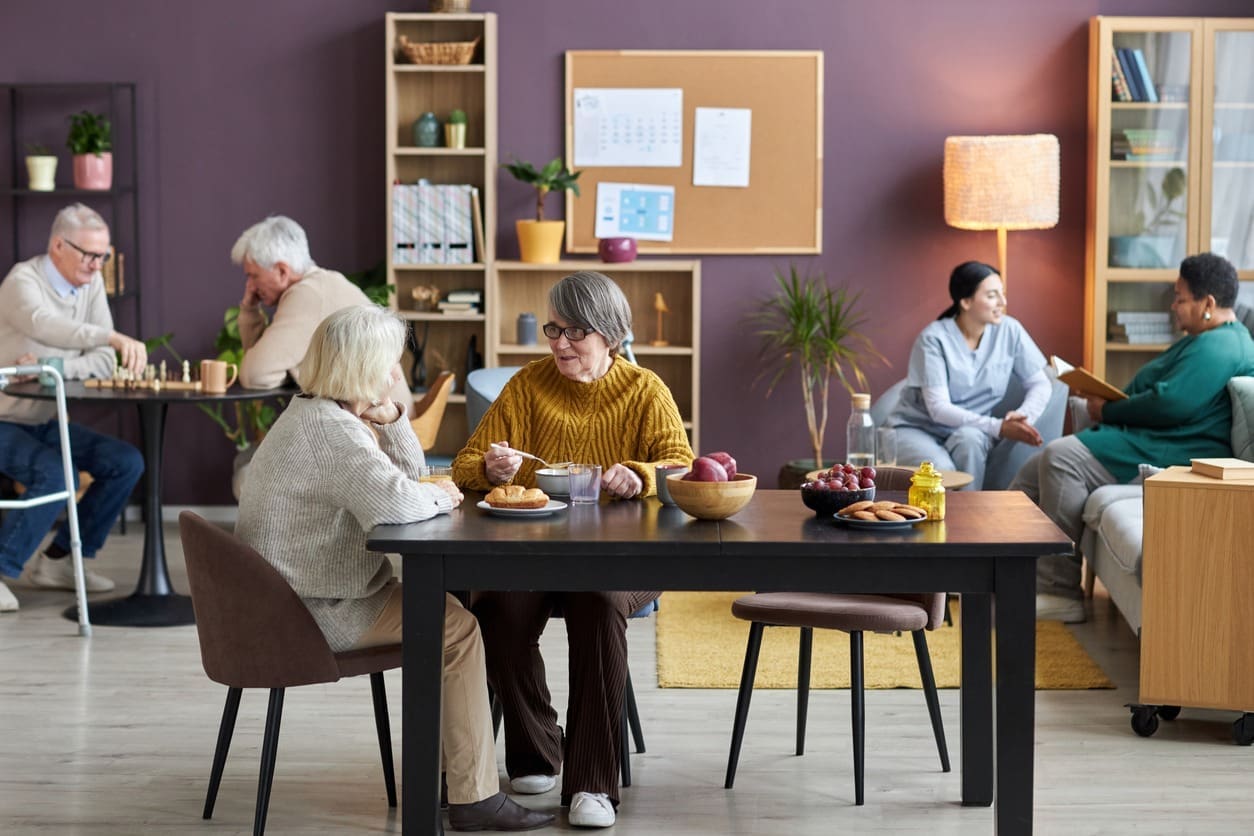 A group of seniors socialize in a cozy, human-centered common room; two women talk at a table with snacks, while others play chess and chat on couches. Shelves and a bulletin board highlight the supportive environment.