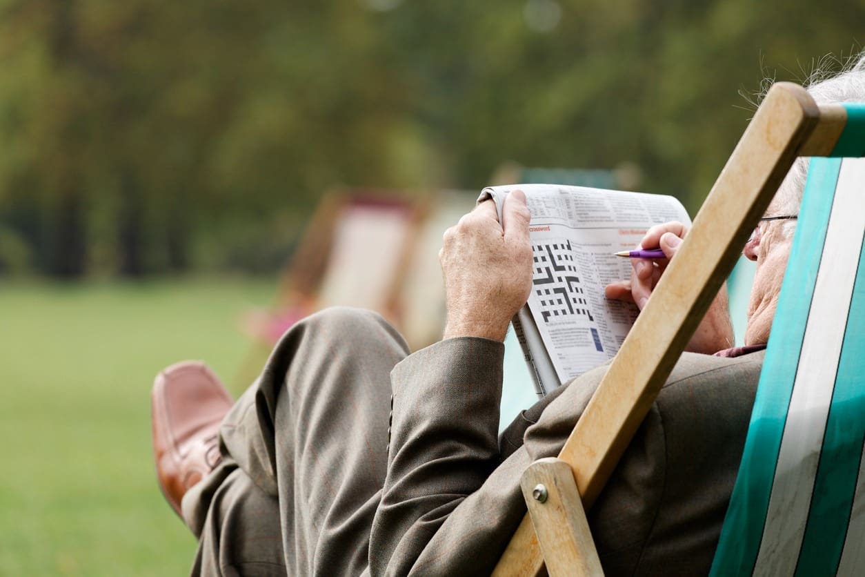 An older man sits on a deck chair outdoors, relaxing on the grass while filling out a crossword puzzleโa meaningful way for families to help spot early symptoms of dementia. He is dressed in a suit and holding a pen, with trees blurred in the background.