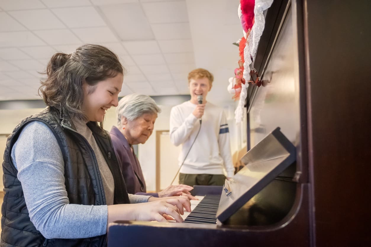 A young woman plays the piano while an older woman sits beside her. In the background, a man stands and sings into a microphone. They all appear to be enjoying the music together in a bright, indoor setting.