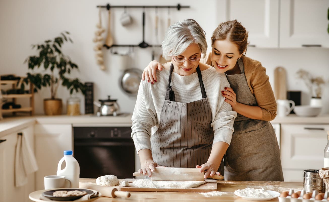 Two women in aprons, one older and one younger, smile while preparing dough together in a kitchen. The older woman rolls out dough on a floured surface, and baking ingredients and utensils are visible on the table.
