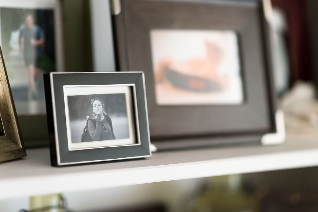 A small black-and-white framed photo of a woman blowing a kiss sits on a white shelf, surrounded by other blurred, larger picture frames in the background.