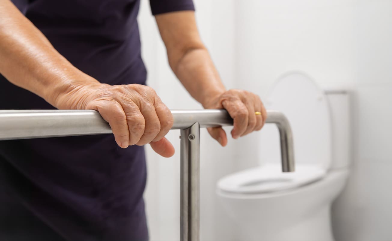 An elderly person holds onto a metal grab bar in a bathroom, with a white toilet in the background, suggesting support and safety for mobility assistance.