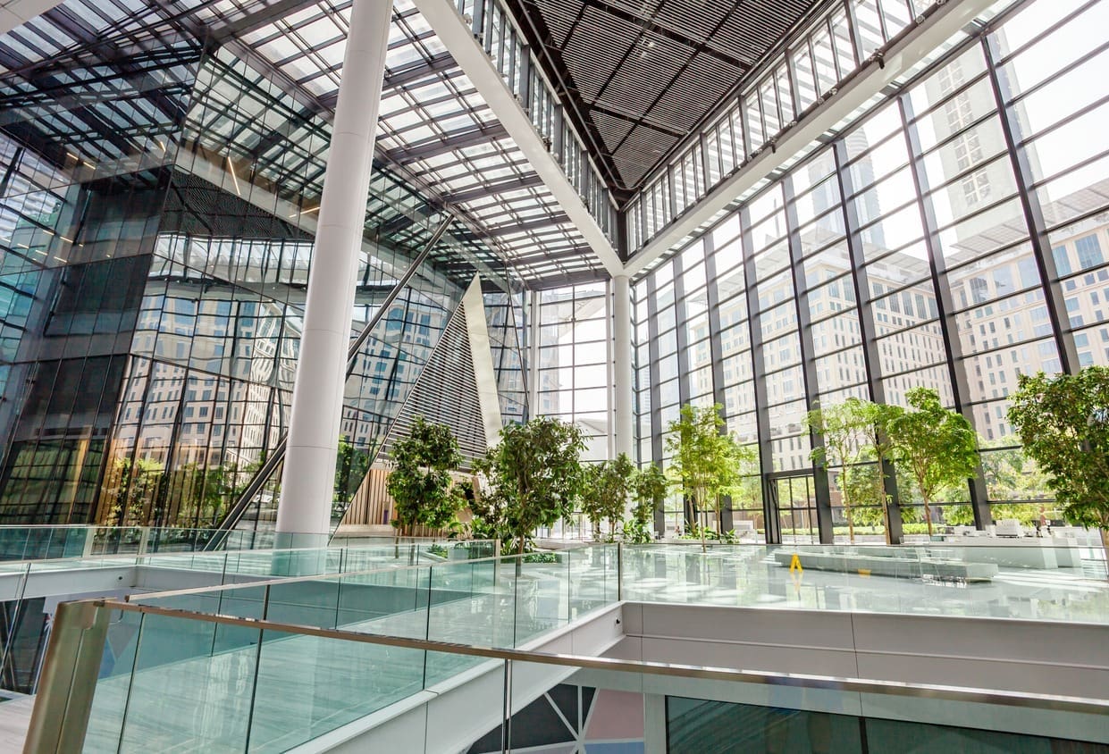 A spacious, modern atrium with high glass walls and ceiling, filled with natural light. Green plants are arranged along walkways, and tall buildings are visible through the windows.
