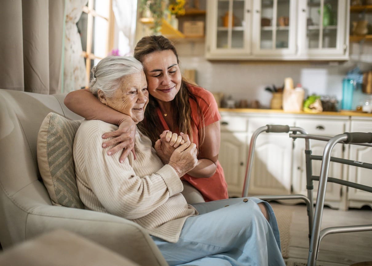 A younger woman hugs an elderly woman on a cozy kitchen couch. Both smile warmly, with a walker beside them, suggesting care and support for early symptoms of dementia in a loving home environment.