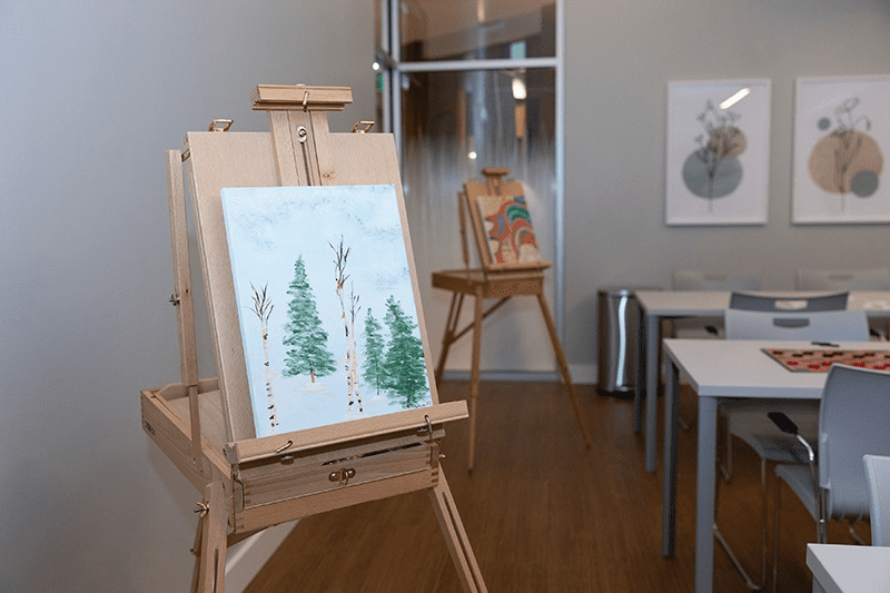 An easel holds a painting of green trees in a bright, modern art room designed with Memory Care Building Design principles, featuring another easel, white chairs, tables, and framed abstract artwork on the wall in the background.