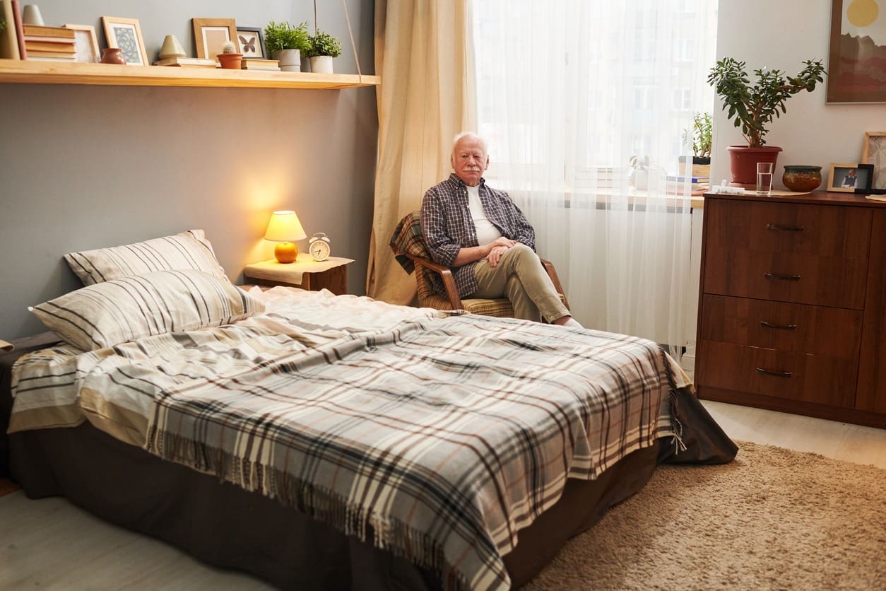 An elderly person sits on a chair next to a neatly made bed in a cozy, sunlit bedroom with wooden furniture, potted plants, and framed photos on shelves.