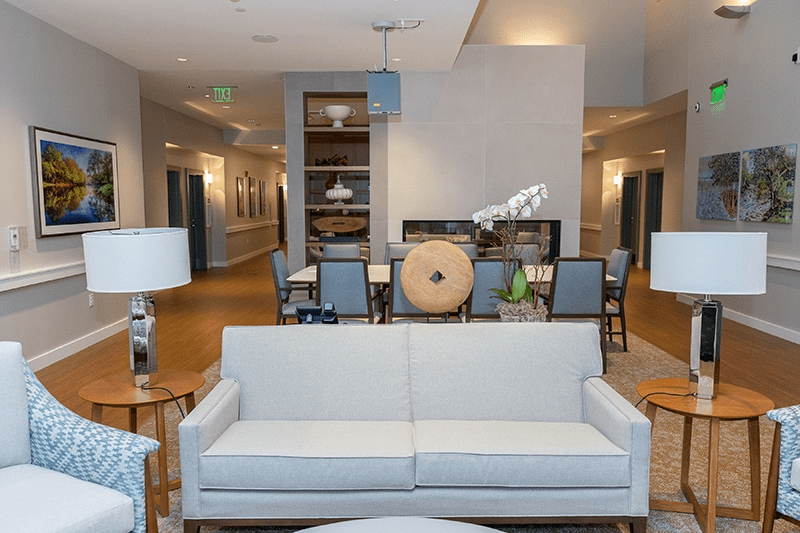 A modern, well-lit lounge area with light-colored sofas, chairs, and tables features neutral decor, wall art, a dining table in the background, and an orchid centerpiece—reflecting thoughtful Memory Care Building Design.