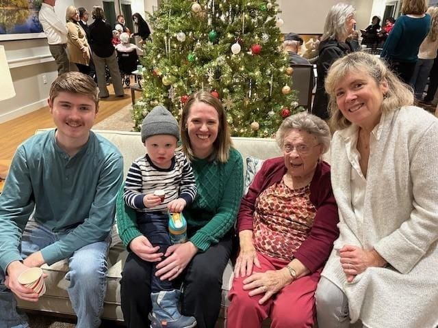 A family sits on a couch in front of a decorated Christmas tree, smiling at the camera; at The Cordwainer, Specialized Memory Care brings joy to gatherings like these, even as people mingle in the background.