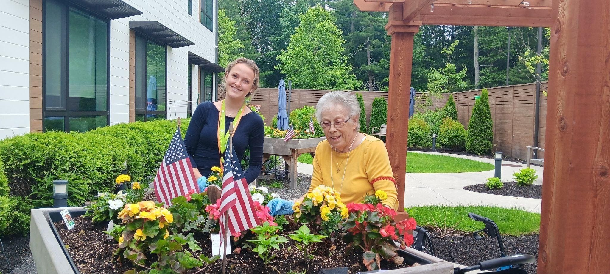 Two women, one younger and one elderly, smile while gardening together at a raised flower bed adorned with colorful flowers and two small American flags, enjoying a sunny day in The Cordwainer’s Dementia Care South Shore MA courtyard.