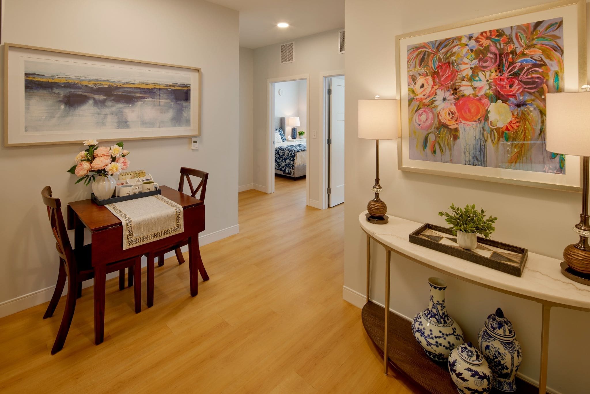 A cozy hallway with wooden floors, a small dining table with flowers, colorful artwork on the walls, and a console table with lamps, a plant, and blue-white vases. Bedroom visible in the background.