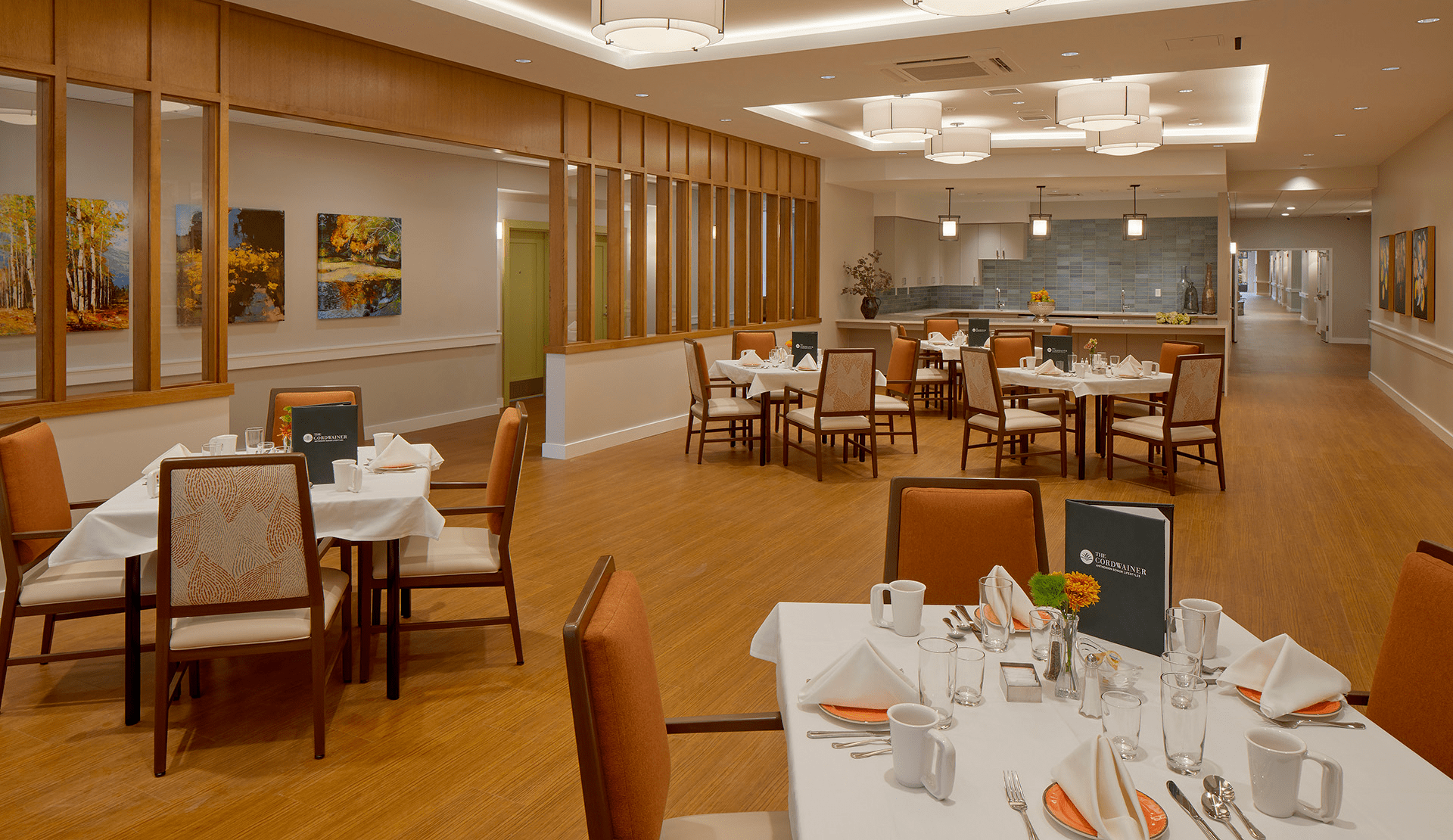 A modern dining room with neatly set tables, wooden chairs, white tablecloths, and menus. Warm lighting, wooden floors, and nature-themed artwork reflect thoughtful Memory Care Building Design for an inviting, comfortable atmosphere.